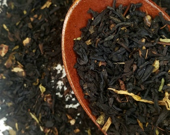 May include: A wooden spoon filled with loose leaf black tea, with a scattering of tea leaves on a white surface.