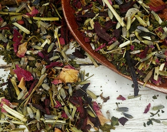 May include: A wooden spoon filled with a blend of dried herbs, flowers, and fruit pieces. The mixture includes green herbs, lavender buds, rose petals, and dried fruit pieces. The spoon is on a white wooden surface.