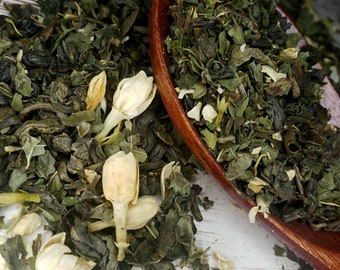 May include: A wooden spoon filled with green tea leaves and white jasmine flowers. The tea is scattered on a white wooden surface.