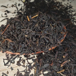 Könnte beinhalten: Lose schwarze Teeblätter in einem braunen Keramiklöffel, mit weiteren Teeblättern, die auf einer hellen Holzoberfläche verstreut sind.