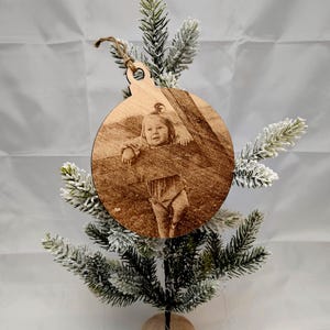 May include: A round wooden ornament with a photo of a young child engraved on it. The ornament is hanging from a small tree.