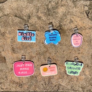 Può includere: Una collezione di ciondoli in acrilico colorati con vari disegni di testo. I ciondoli includono frasi come "Kindle Girl", "Currently Escaping Reality" e "Just One More Page". Ogni ciondolo ha una clip trasparente.