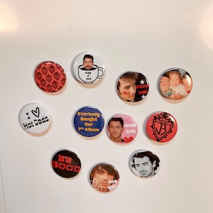 May include: A collection of round pinback buttons with various designs and text. Some buttons feature images of people, while others have phrases like "I love Hot Dads" and "Be Good." Colors include red, white, blue, and black.