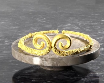 May include: A gold-colored necklace with intricate scrollwork details. The necklace is displayed on a circular, light brown stand.