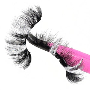 Puede incluir: Dos pares de pestañas postizas con una banda de purpurina plateada en cada pestaña. Las pestañas están en una pinza rosa.