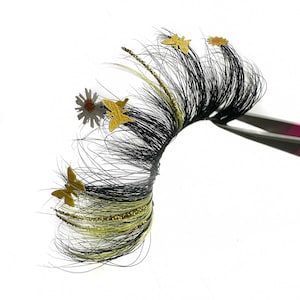 May include: A single false eyelash with black and yellow strands, accented with gold butterfly and daisy embellishments. The lash is held by pink-handled tweezers against a white background. The lash has a dramatic, full appearance.