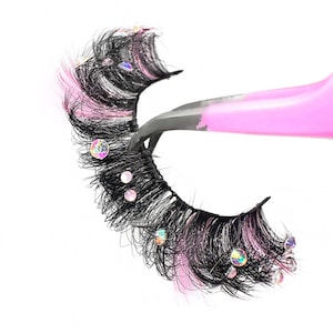 May include: A pair of false eyelashes with pink and black fibres and iridescent rhinestones. The eyelashes are held by a pink eyelash applicator.