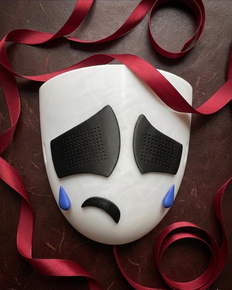May include: A white mask with black eye coverings and a black curved mouth, with blue teardrops. Red ribbon is draped around the mask, creating a decorative display. The mask has a sad expression.
