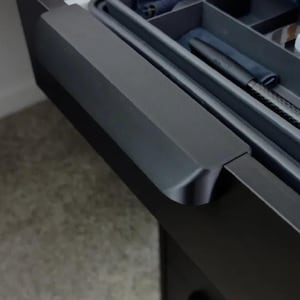 May include: A partially open, dark gray drawer with a built-in organizer. The drawer contains various items, including a pen and a cloth. The drawer is part of a larger cabinet with a wooden top.