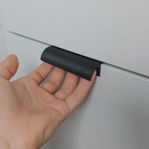 Könnte beinhalten: Ein schwarzer Schrankgriff ist an einer weißen Schranktür montiert. Der Griff hat eine gebogene, rechteckige Form. Die Schranktür ist hellgrau.