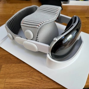 May include: A white VR headset with a grey fabric headband is resting on a white charging stand. The headset has a black lens and a white charging cable is connected to the stand.