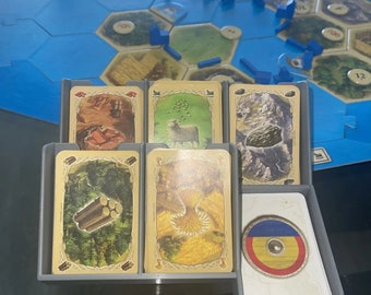 May include: A board game with a blue and tan game board and gray card holders. The cards feature illustrations of resources like wood, sheep, and wheat. A stack of cards with a circular emblem is also visible.