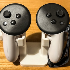 Könnte beinhalten: Ein weißer VR-Controller-Ladeständer mit zwei schwarzen VR-Controllern. Jeder Controller hat ein kreisförmiges Tastenfeld mit verschiedenen Tasten, darunter A, B, X, Y und einen Joystick.