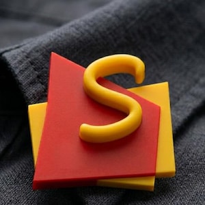 Op de afbeelding: Een gele letter "S" ligt bovenop een rode driehoek en een gele vierkant. De geometrische vormen zijn op elkaar gelegd. De achtergrond is een donkergrijze stof.