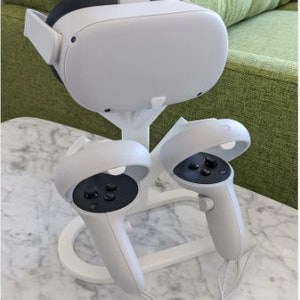 Może przedstawiać: Biały stojak na zestaw VR z dwoma białymi kontrolerami. Stojak ma białą podstawę i białe, Y-kształtne wsparcie dla zestawu. Kontrolery są przymocowane do podstawy białymi klipsami.