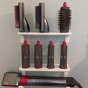May include: A Dyson hair styling set, including a hair dryer and multiple styling attachments, displayed on a white wall-mounted shelf. The attachments are various shapes and sizes, with gray and pink accents. The hair dryer is gray with pink details.