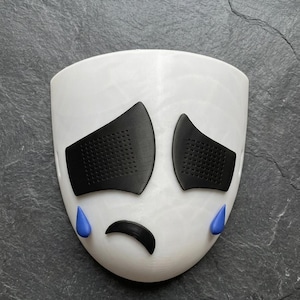 May include: A white mask with black eye coverings and a curved black mouth. Blue teardrops are positioned below the eyes. The mask is designed to resemble a sad face.