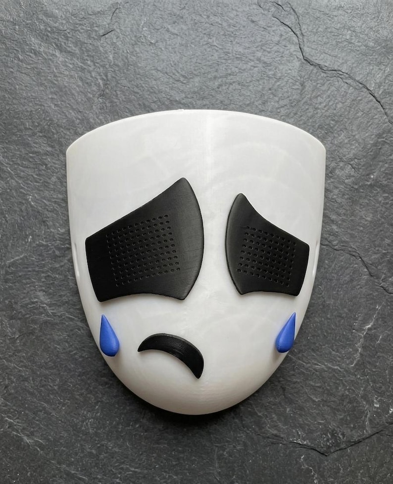 May include: A white mask with black eye coverings and a curved black mouth. Blue teardrops are positioned below the eyes. The mask is designed to resemble a sad face.