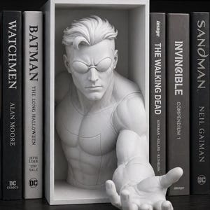 Op de afbeelding: Een witte sculptuur van een mannelijke figuur met een gespierde torso en uitgestrekte hand, geplaatst in een wit rechthoekig frame. De sculptuur staat tussen verschillende zwarte boeken met witte letters, waaronder "Watchmen" en "Batman".