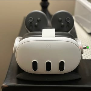 Op de afbeelding: Witte virtual reality-headset met zwarte controllers en een groen lampje aan de zijkant. De headset heeft drie zwarte sensoren aan de voorkant en een oneindigheids symbool logo.
