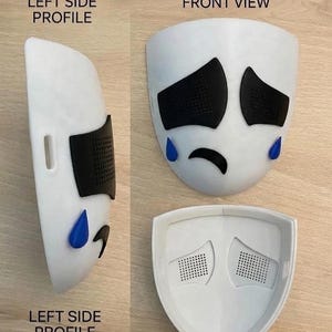 May include: A white mask with black eye and eyebrow shapes, and blue teardrops. The mask has a sad face design. The image shows the front, side, and rear views of the mask. The text "LEFT SIDE PROFILE", "FRONT VIEW", and "REAR VIEW" are visible.