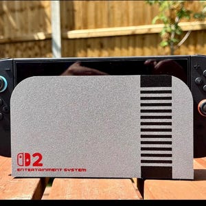 May include: A black Nintendo Switch console with a gray cover featuring a design reminiscent of the classic Nintendo Entertainment System. The cover has the text "Switch 2 Entertainment System" in red. The console is on a wooden surface.
