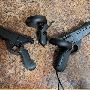 May include: Two black VR controllers with gun-shaped attachments. The controllers are disassembled and lying on a brown work surface.