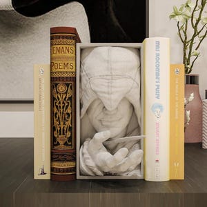 May include: A still-life arrangement featuring several books, including one titled "HEMANS POEMS" with ornate gold lettering. A framed sculpture of a face and hand is centered. Vases and decorative items complete the scene.