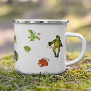 May include: White enamel mug with a variety of frog illustrations. The mug has a silver rim and handle. The frogs are depicted in various poses and colors, including green, red, and yellow.