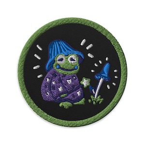 Könnte beinhalten: Gestickter Aufnäher mit einem grünen Frosch, der einen blauen Hut und einen violetten Schlafanzug trägt und einen leuchtenden Stern hält. Der Frosch steht neben einem blauen Pilz mit einem weißen Stiel. Der Aufnäher hat einen grünen Rand und ist auf einem schwarzen Hintergrund.