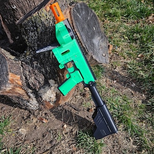 May include: A green and black toy gun with an orange barrel. The gun is lying on a tree stump in a grassy area.
