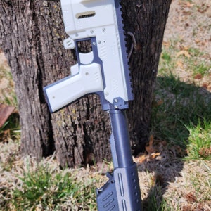 May include: A white and black toy gun with an orange tip. The gun is made of plastic and has a realistic design. It is sitting on a tree stump in a grassy area.