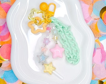 Może przedstawiać: Wisiorek z zielonego łańcuszka w pastelowych kolorach z gwiazdą, lizakiem i kwiatem. Wisiorek leży na białym talerzu otoczonym kolorowym konfetti.