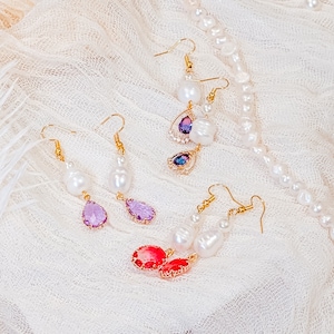 May include: Three pairs of pearl and gemstone earrings. The earrings feature a teardrop-shaped pearl with a gemstone accent. The gemstones are purple and red.