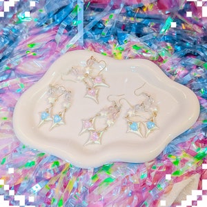 May include: Four pairs of gold star-shaped earrings with pink and blue crystal accents. The earrings are displayed on a white ceramic dish.