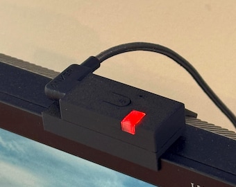 May include: A black rectangular webcam privacy cover with a red LED indicator light attached to the top edge of a monitor. The device features a small button and a black cable extending from the side, with the text VA2323 visible on the monitor bezel.