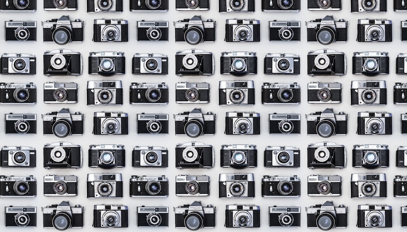 May include: A wall covered in a grid pattern of vintage silver and black 35mm film cameras.