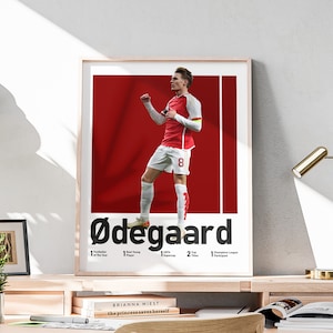 May include: A red and white poster with a photo of a soccer player celebrating a goal. The player is wearing a red and white jersey with the number 8 on it. The poster has the text "Ødegaard" in black letters. The poster also has text about the player's accomplishments, including "1 Footballer of the Year", "1 Best Young Player", "1 UEFA Super Cup", "2 Cup Titles", and "1 Champions League Participant".