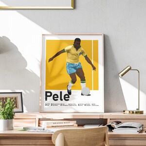 May include: A framed print depicting Pel&eacute; in a yellow and blue football kit. The print has a yellow background with white vertical lines and text detailing his accomplishments. The print is displayed on a wooden desk, with a gold lamp.
