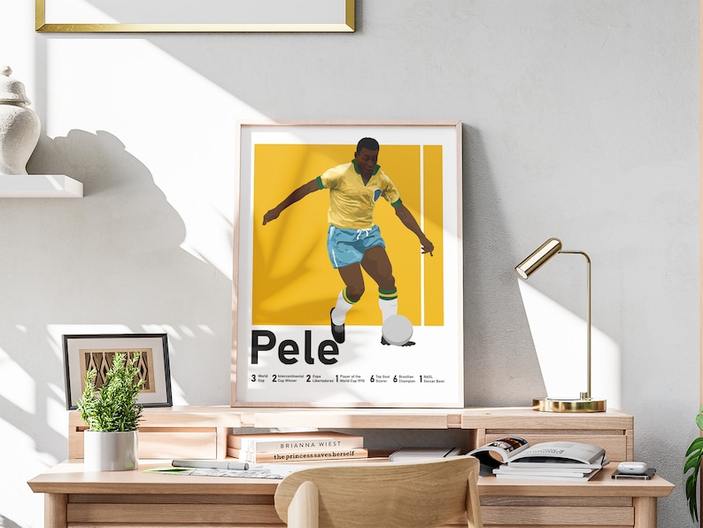 May include: A framed print depicting Pel&eacute; in a yellow and blue football kit. The print has a yellow background with white vertical lines and text detailing his accomplishments. The print is displayed on a wooden desk, with a gold lamp.