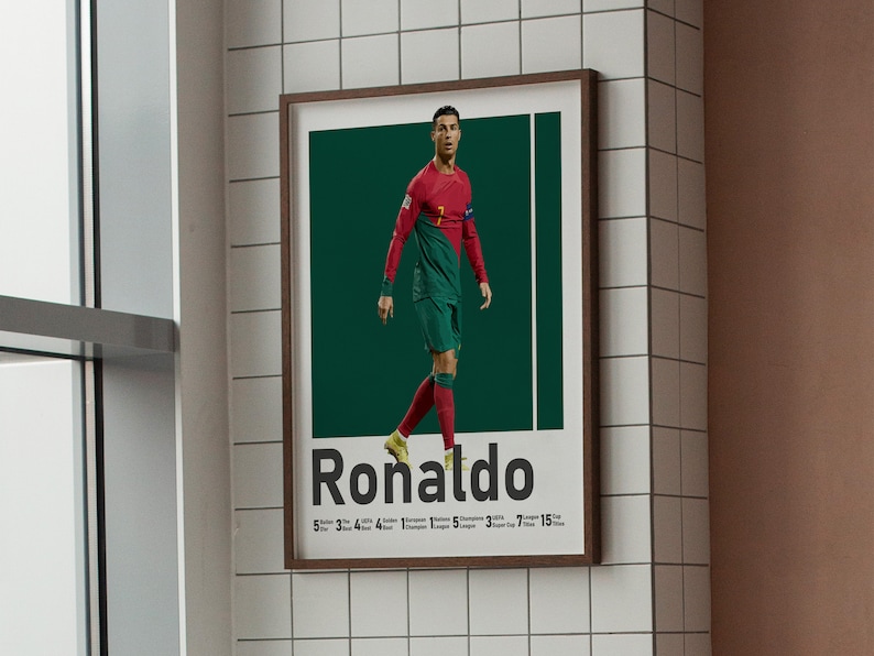 May include: A framed poster of Cristiano Ronaldo in a red and green football kit against a dark green backdrop. The name "Ronaldo" is in bold text below the image, with statistics beneath. The poster is displayed on a white tiled wall, with a brown frame.