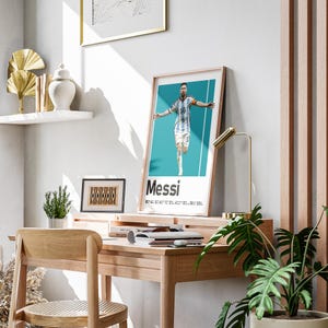 May include: A framed poster featuring Lionel Messi in his Argentina national team jersey, with the name "Messi" in bold white text. The poster is displayed on a wooden desk, alongside a gold desk lamp and a small potted plant.