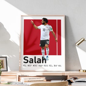 K&ouml;nnte beinhalten: Ein rotes und wei&szlig;es Poster mit einem Fu&szlig;ballspieler in einem wei&szlig;en Trikot mit der Nummer 10. Der Spieler ist im Laufschritt mit erhobenem rechten Arm. Das Poster tr&auml;gt den Text "Salah" in gro&szlig;en schwarzen Buchstaben unten. Das Poster enth&auml;lt auch eine Liste der Leistungen des Spielers, darunter "FIFA Puskas Award", "Champions League", "Premier League" und "Africa Cup of Nations".