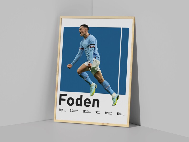 May include: A framed poster featuring a footballer in mid-air, celebrating a goal. The player is wearing a light blue shirt with white accents. The poster has a blue background and the player's name, "Foden", is printed in bold black letters at the bottom. The poster also lists the player's achievements, including 1 Super Cup, 1 Champions League, 5 English Champions, 9 Cup Titles, 1 World Cup Participant, and 1 Euro Participant.