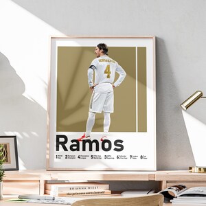 May include: A poster featuring a white and gold illustration of a soccer player wearing a white jersey with the number 4 on the back. The player's name, "Ramos", is printed in large, bold black letters below the illustration. The poster also includes a list of the player's accomplishments, including the number of times he has won the Champions League, World Cup, and other titles.
