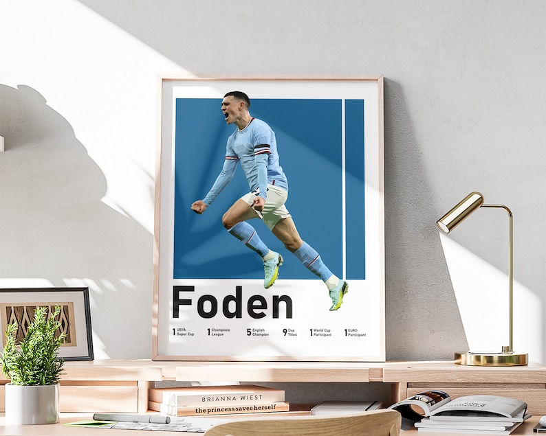 May include: A poster featuring a football player in mid-air, celebrating a goal. The player is wearing a light blue jersey with white accents and white shorts. The background is a solid blue. The poster has the player's name "Foden" in bold black text and a list of his achievements.