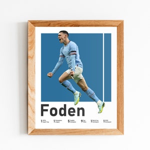 May include: A framed poster of a footballer in mid-air, celebrating a goal. The player is wearing a light blue shirt with white accents and black shorts. The poster has a blue background and the player's name, "Foden", is printed in bold black letters at the bottom. The poster also lists the player's achievements, including 1 Super Cup, 1 Champions League, 5 English Championships, 9 Cup Titles, 1 World Cup Participant, and 1 EURO Participant.