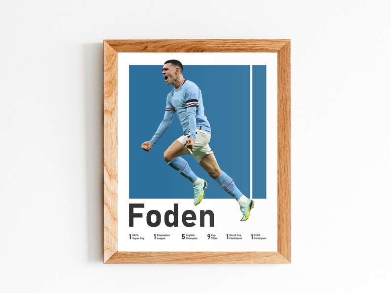 May include: A framed poster of a footballer in mid-air, celebrating a goal. The player is wearing a light blue shirt with white accents and black shorts. The poster has a blue background and the player's name, "Foden", is printed in bold black letters at the bottom. The poster also lists the player's achievements, including 1 Super Cup, 1 Champions League, 5 English Championships, 9 Cup Titles, 1 World Cup Participant, and 1 EURO Participant.