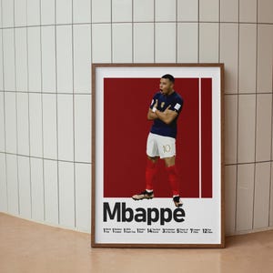 Puede incluir: Un p&oacute;ster enmarcado que muestra a un futbolista con una camiseta azul oscuro y pantalones cortos blancos con el n&uacute;mero 10, sobre un fondo rojo intenso. El texto Mbapp&eacute; aparece debajo, seguido de una lista de logros y estad&iacute;sticas.