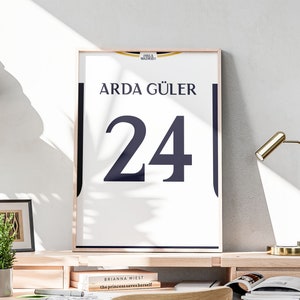 May include: A white framed poster with a black and white design. The poster features the text "Real Madrid" and "Arda Güler" with the number "24" in large black font.